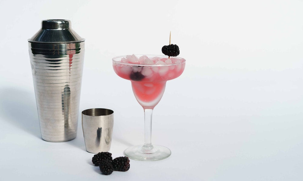 Bramble Cocktail im Glas mit Brombeeren garniert neben einem Shaker auf weißem Hintergrund